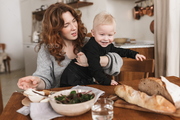 Young beautiful woman with red hair in knitted sweater dreamily feeding her little smiling son - Stock Photo - Images
