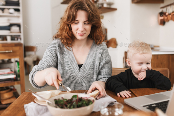 Redhead woman in sweater dreamily cooking while her little son happily watching cartoons on laptop - Stock Photo - Images