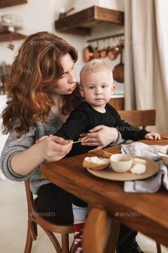 Young redhead woman in knitted sweater sitting at the table with food dreamily feeding her baby - Stock Photo - Images