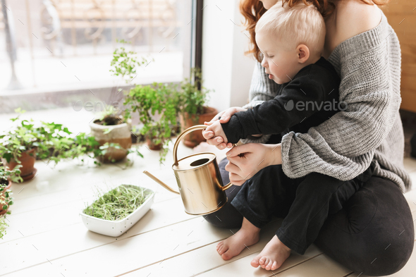 Young mother sitting on floor with her little son watering green plants around near big window