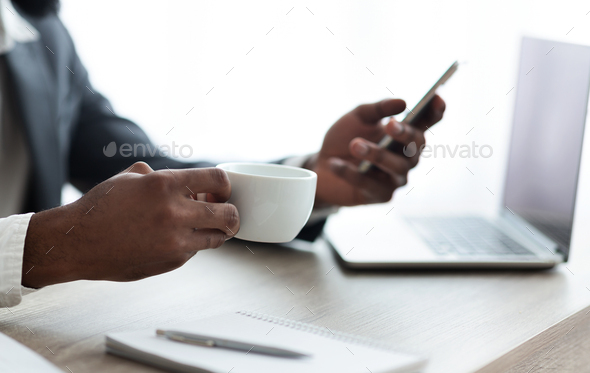 Black businessman drinking coffee and using smartphone at workplace