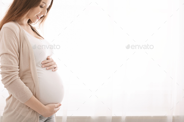 Happy pregnant woman touching her belly near window