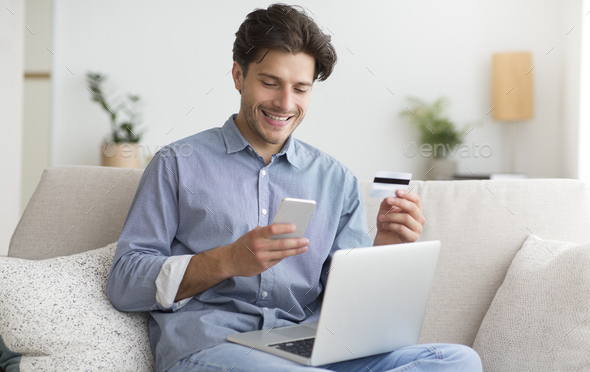 Guy Using Phone, Credit Card And Laptop Providing Payment Indoor