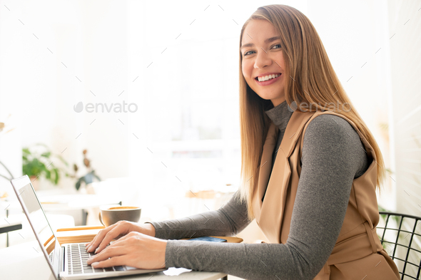 Young elegant businesswoman looking at you with toothy smile while typing