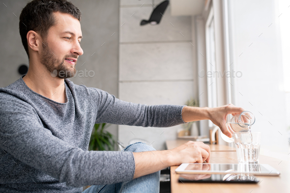 Casual man sitting by window in cafe and pouring water from bottle into glass