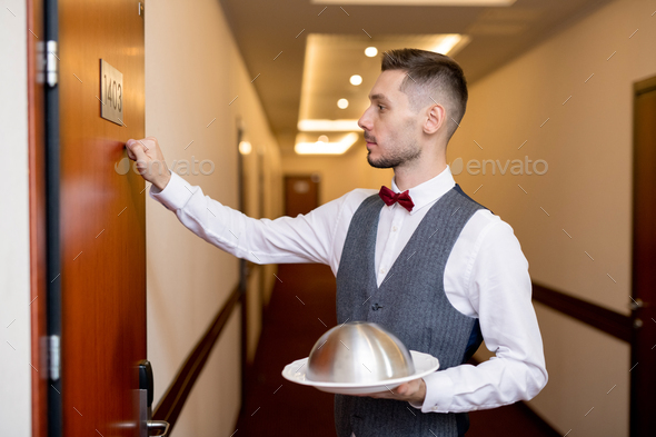 Young elegant waiter with food on cloche for one of guests knocking on the door