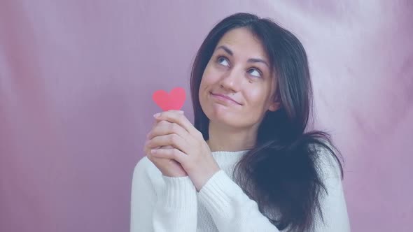 Smiling Funny Young Brunette in a White Sweater Smiles Blinks and Waving Red Hearts on a Pink