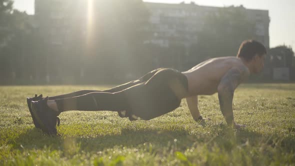 Young Caucasian Sportsman Pushing Up in Sunrays. Side View Portrait of Strong Tattooed Man alt
