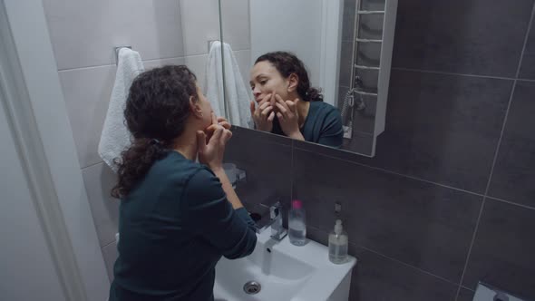 Woman Removing Pimple From Face Looking at Mirror in Bathroom Home alt