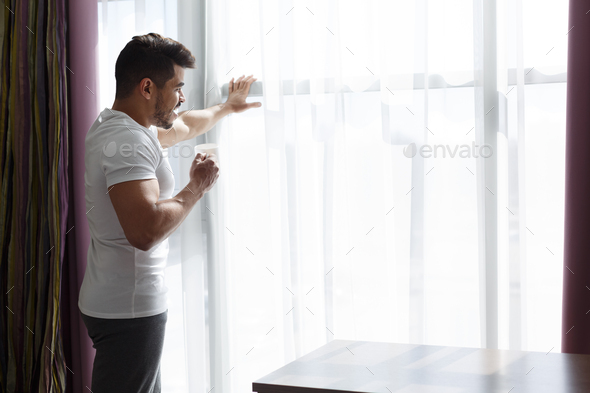 Young guy staying next to window, drinking morning coffee
