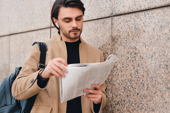 Young confident stylish man in beige coat with backpack reading newspaper outdoor
