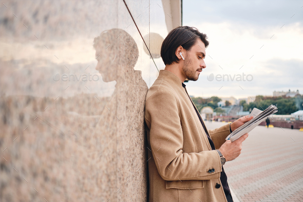 Side view of young handsome stylish man in beige coat reading newspaper outdoor