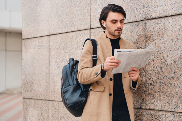 Young attractive casual man in beige coat with backpack thoughtfully reading newspaper outdoor