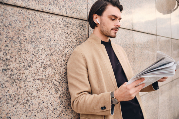 Young handsome stylish man in beige coat thoughtfully reading newspaper outdoor