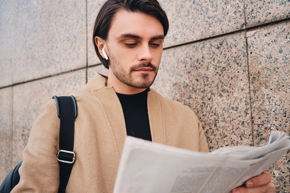 Young confident casual brunette man in beige coat thoughtfully reading newspaper outdoor