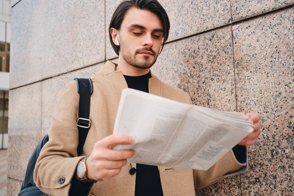 Portrait of young wistful brunette man in beige coat reading newspaper outdoor