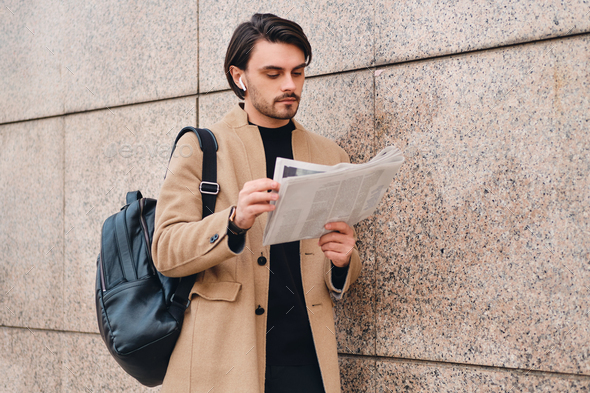 Young thoughtful casual man in beige coat with backpack reading newspaper outdoor