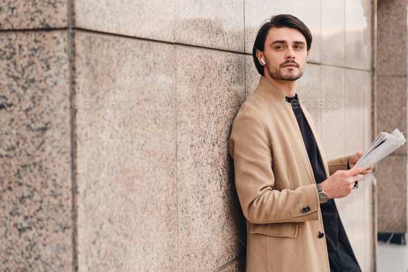 Attractive stylish man in beige coat thoughtfully looking away standing with newspaper outdoor