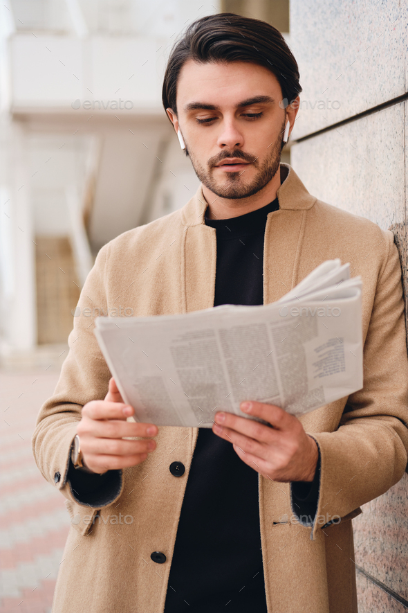 Young attractive casual man in beige coat reading newspaper outdoor