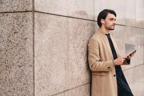 Young handsome stylish man in beige coat with wireless earphones reading newspaper outdoor