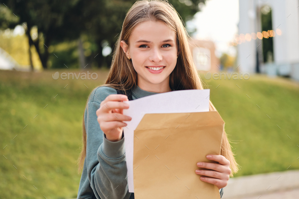Cheerful student girl opening envelope with exams results happily looking in camera in city park
