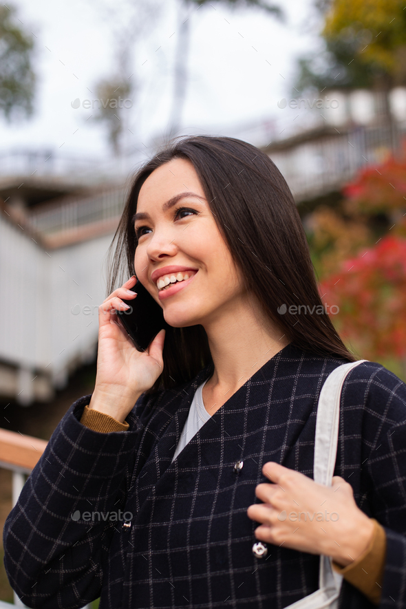 Side view of young smiling casual woman in coat happily talking on cellphone in city park
