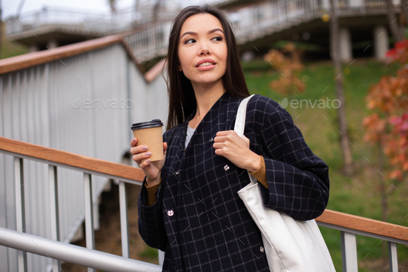 Young attractive casual woman in coat with coffee to go thoughtfully looking away in city park