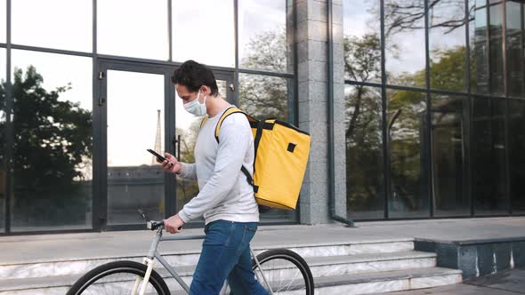 Courier with Yellow Backpack and Protective Mask Walking with Bicycle in City Center and Using alt