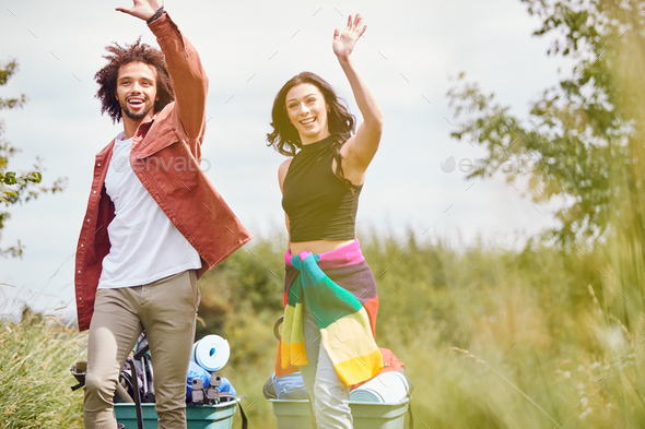 Young Waving Couple Pulling Trolley Filled With Camping Equipment Through Field To Music Festival