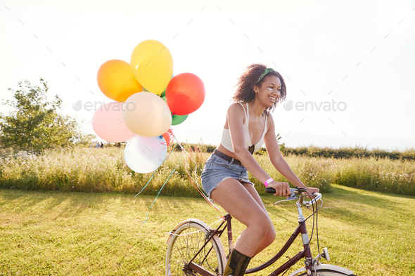 Young Woman Riding Bicycle Decorated With Balloons Through Countryside Against Flaring Sun