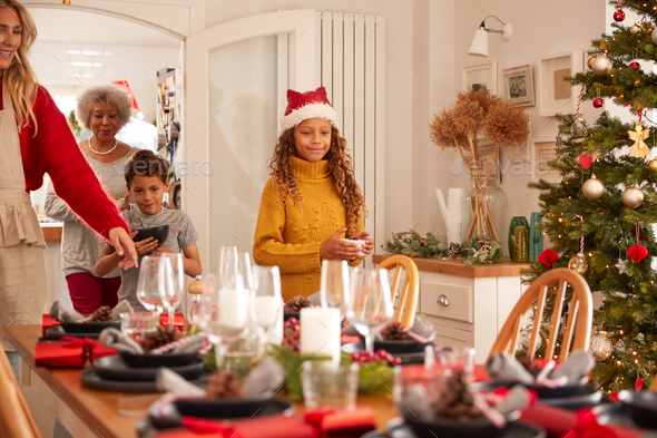 Multi-Generation Family In Dining Room Putting Christmas Meal On Decorated Table