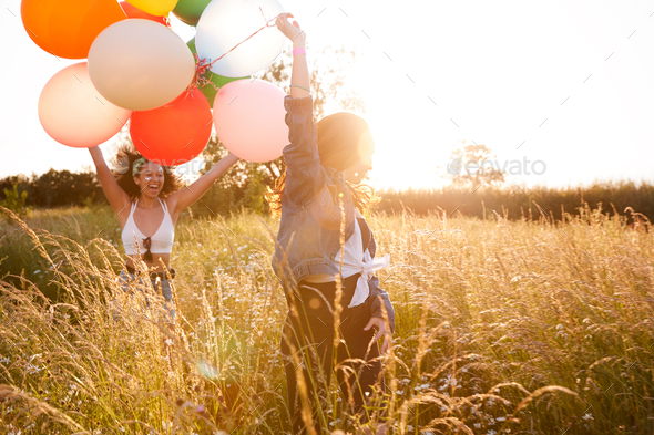 Two Female Friends Camping At Music Festival Running Through Field With Balloons Against Flaring Sun