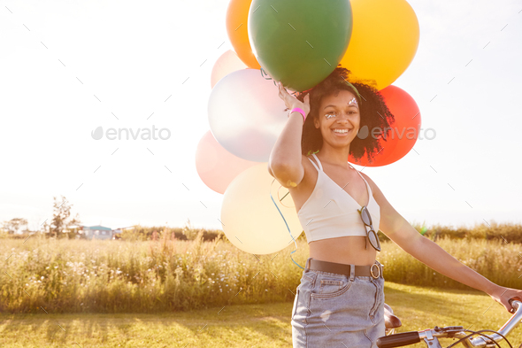 Portrait Of Woman Riding Bicycle Carrying Balloons Through Countryside Against Flaring Sun