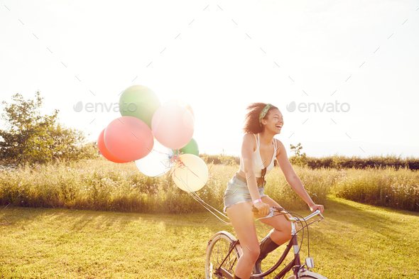 Young Woman Riding Bicycle Decorated With Balloons Through Countryside Against Flaring Sun