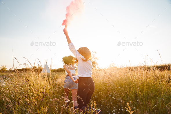 Rear View Of Two Female Friends Running Through Field With Smoke Flare