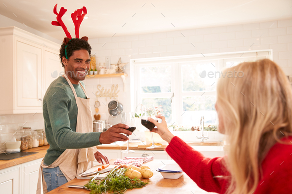 Couple Wearing Fancy Dress Antlers Making A Toast Whilst Preparing Dinner On Christmas Day