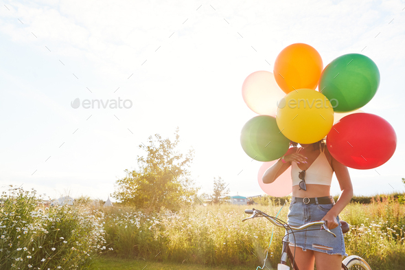 Young Woman With Bicycle Hidden By Balloons Rides Through Countryside Against Flaring Sun