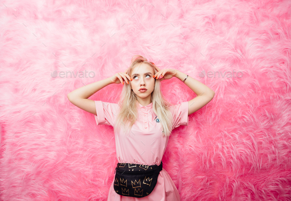 Young beautiful blonde girl dressed in fashion pink dress poses on the  background of pink fur wall Stock Photo by leikapro