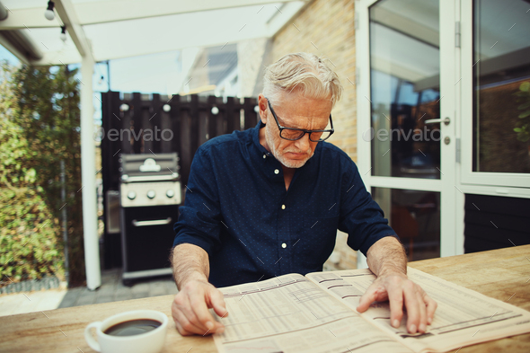 Stern senior man reading a financial newspaper at home - Stock Photo - Images