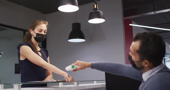 Caucasian woman in face mask has temperature checked by male colleague at office reception desk alt
