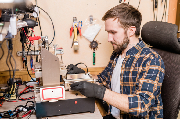 Bearded repairman in gloves sitting in front of equipment for gadget repair