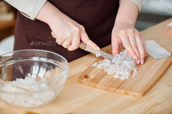 Hands of young craftswoman with knife cutting transparent soap mass on board