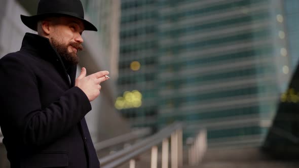 Strange Man in Black Coat and Hat is Smoking Cigarette and Waiting Someone on Street in Autumn Day alt