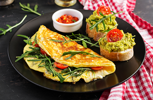 Omelette with tomatoes, cheese, green arugula and toasts with avocado cream on black plate. - Stock Photo - Images