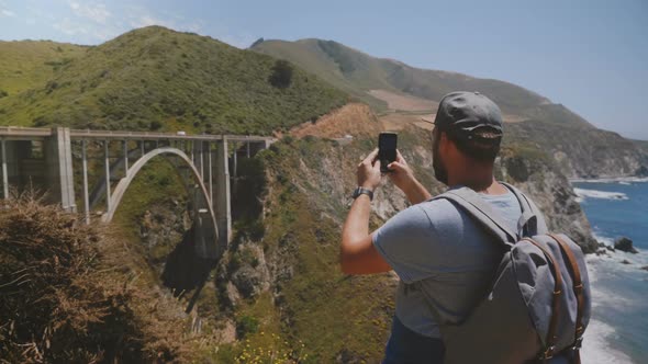 Happy Middle Aged Blogger Tourist Man with Backpack Takes Panoramic Smartphone Video of Iconic Bixby alt