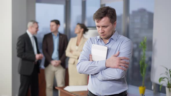 Portrait of Stressed Overworked Man Holding Tablet Looking Back at Blurred Colleagues Talking alt