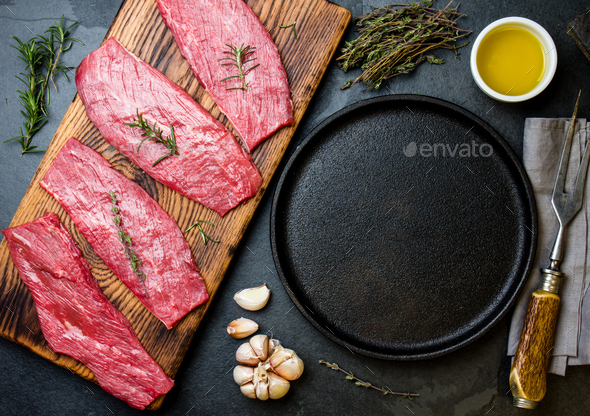 Beef tenderloin in cast iron pan on wooden board, spices, herbs, oil on slate gray background - Stock Photo - Images