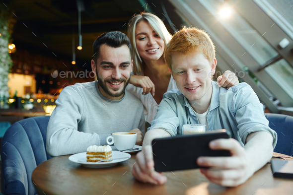 Two happy guys and pretty blonde girl making selfie on smartphone in cafe