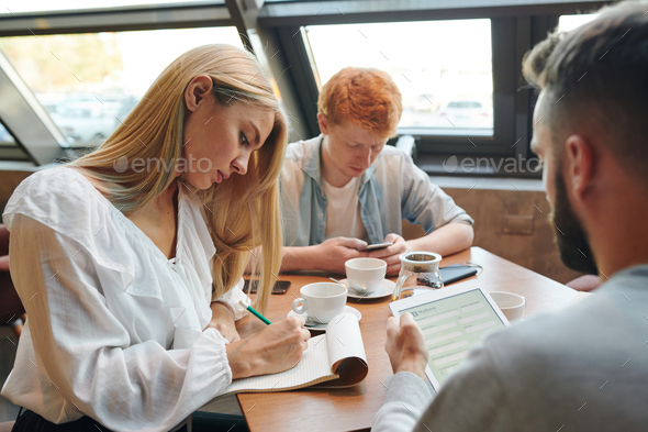 Two clever guys using gadgets while blonde girl making notes in notepad in cafe
