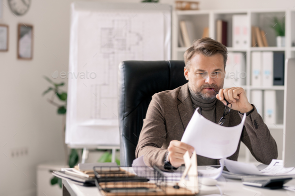 Pensive mature architect looking at sketch on paper while sitting in armchair